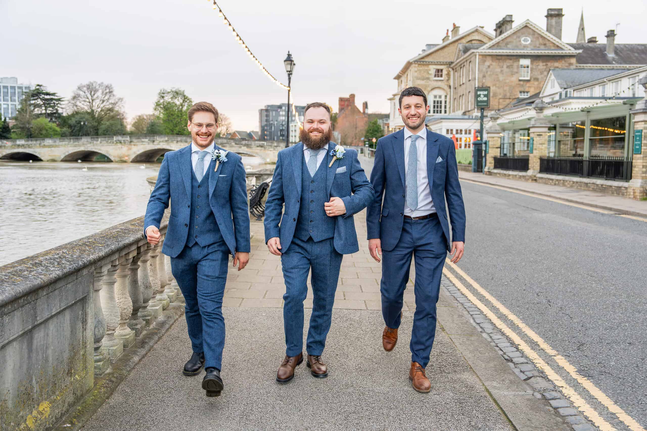 Groom and groomsmen walking along the river promenade at Bedford Hotel & Spa, Bedfordshire, white flower boutonnieres on their lapels. The man in the middle, donning a waistcoat and beard, exudes confidence. They appear cheerful against the backdrop of an old building and bridge near the Bedford Swan Hotel and Spa. The sky is overcast.