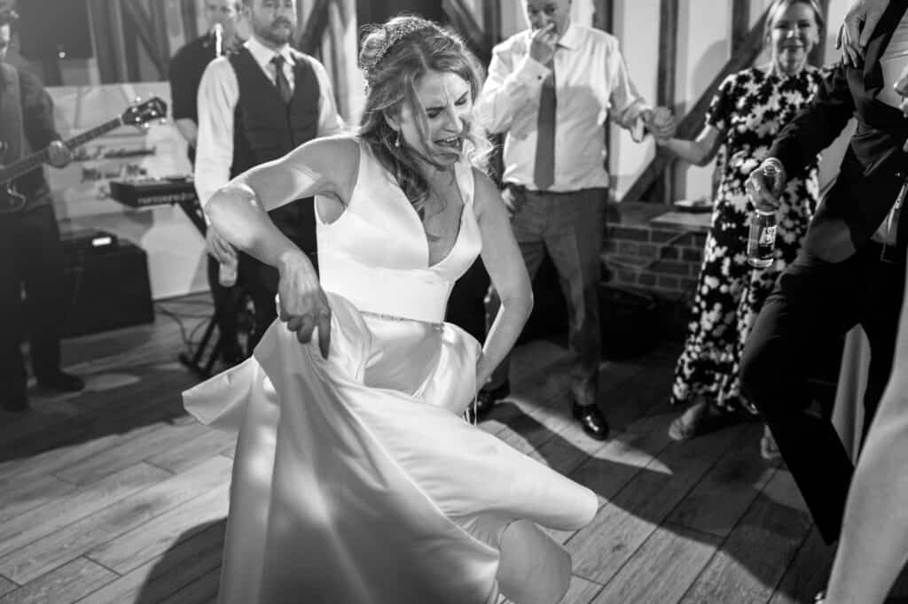 A bride in a white dress energetically dances at her wedding reception at milling barn, hertfordshire, lifting her dress slightly. People around her are dancing, smiling, and holding hands. In the background, a man plays the guitar, adding to the lively atmosphere. Black and white image.