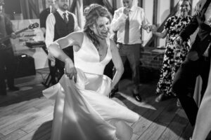 A bride in a white dress energetically dances at her wedding reception at milling barn, hertfordshire, lifting her dress slightly. People around her are dancing, smiling, and holding hands. In the background, a man plays the guitar, adding to the lively atmosphere. Black and white image.