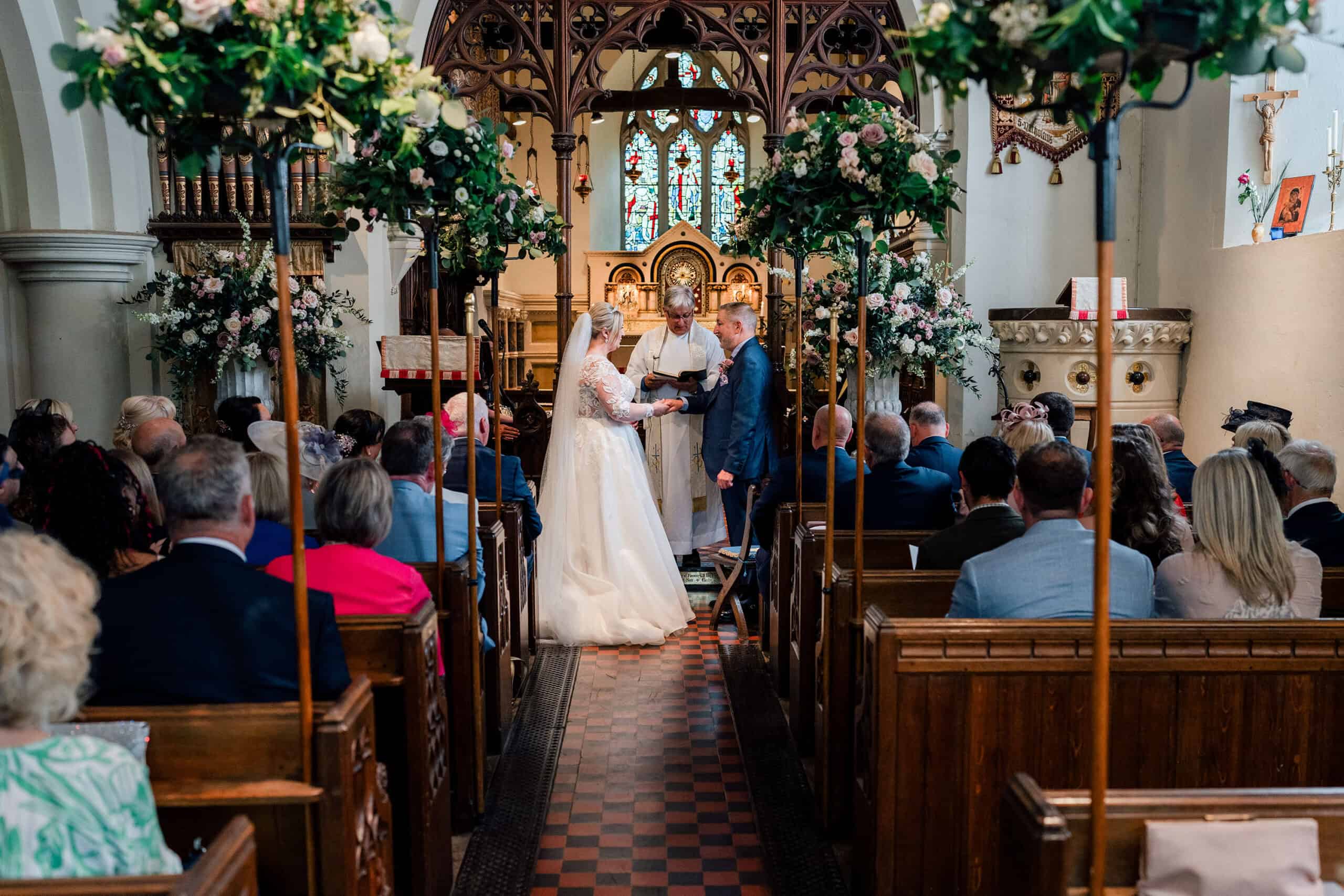 A couple stands at the altar in a church at Hedsor house, Buckinghamshire, holding hands, as a vicar oversees their wedding ceremony. Guests are seated in wooden pews on either side of the aisle, which is adorned with floral arrangements. Stained glass windows and religious symbols are visible in the background.