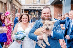 A bride and groom, both smiling, walking through confetti. The groom, who has a beard, is holding a small tan Cairn terrier. The bride carries a bouquet with blue and white flowers. Guests around them are throwing white confetti, and some are also smiling and clapping.
