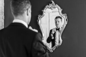 The groom, adjusting his bow tie in front of an ornate mirror. He is seen from the back, with the reflection showing his focused expression. The background features a decorative wall and a door. The photo is in black and white.