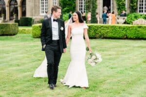 A bride and groom walk hand in hand on a lush green lawn at the back of Hedsor House, Buckinghamshire. The bride wears a white, off-shoulder wedding gown and holds a bouquet of white roses. The groom is dressed in a black tuxedo. They smile at each other and guests are seen blurred in the background drinking.