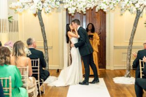 A bride and groom share a kiss under an arch of white flowers, while a smiling officiant looks on. Guests seated in rows on both sides applaud. The ceremony takes place in a warmly lit room with wooden floors and cream-colored walls at Hedsor House, Buckinghamshire.
