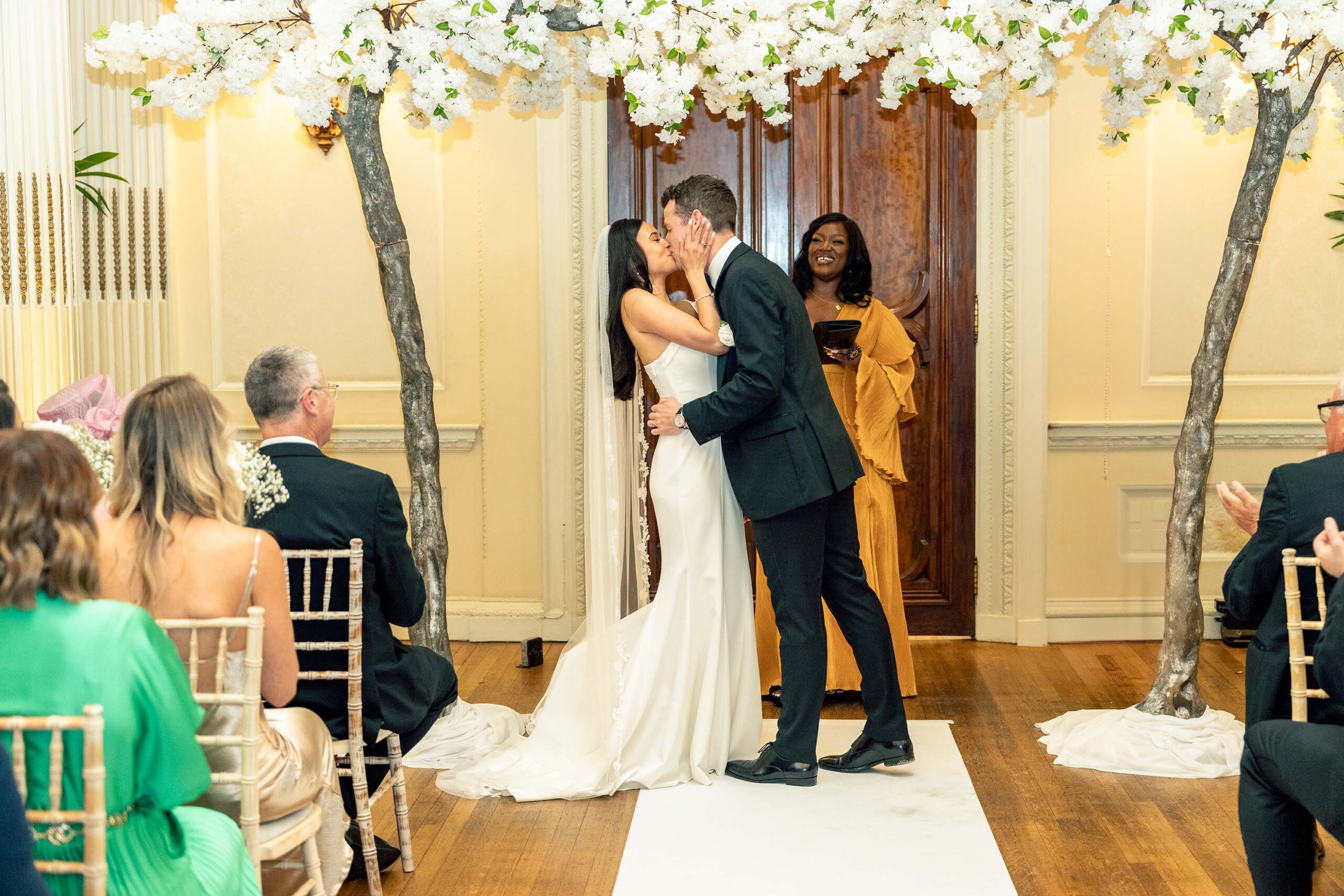 A bride and groom share a kiss under an arch of white flowers, while a smiling officiant looks on. Guests seated in rows on both sides applaud. The ceremony takes place in a warmly lit room with wooden floors and cream-colored walls at Hedsor House, Buckinghamshire.