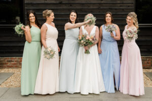 A bride in a white dress stands with five bridesmaids, who are wearing dresses in pastel colors, in front of a dark wooden wall at Milling Barn, hertfordshire. One bridesmaid playfully holds a bouquet of flowers in front of the bride’s face, causing both to laugh.