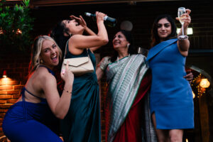 A group of four women celebrate outdoors at night. One woman in green is drinking from a bottle, another in a blue dress is joyfully pointing, a woman in a sari smiles, and another in a blue dress holds up a glass of champagne. They all look very happy.