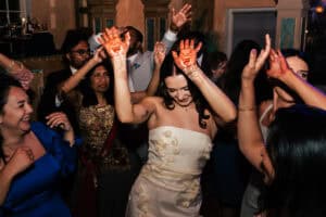 A woman in a white dress, with henna on her hands, dances joyfully with a group of people iat her wedding reception. Others are smiling and dancing around her, wearing colorful attire.