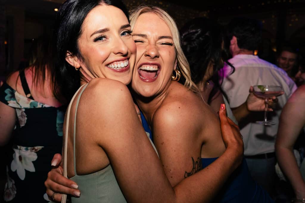 Two women, one with dark hair and one with blonde hair, are enjoying themselves at a wedding at Trading Boundaries, West Sussex. They are hugging, smiling, and laughing. The background is filled with other people, some with drinks in hand, creating a lively and festive atmosphere.