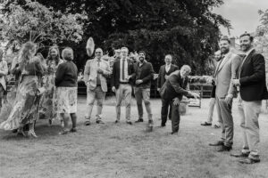 A black-and-white photo of a group of people outdoors at a wedding at Sandon Manor, Hertfordshire. They are throwing bean bags at stacks of tin cans at the garden games. They are laughing and interacting with each other. One person is mid-action, appearing to throw a bean bag, and everyone looks in that direction.