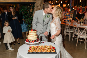 A bride and groom share a kiss while cutting a two-tiered, semi-naked wedding cake decorated with berries at Sandon Manor, Hertfordshire. The bride's white lace dress is complemented by the groom's light gray suit. Guests, including a young girl, observe in the background.