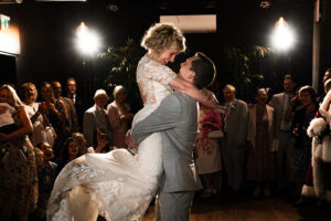 A bride and groom share a joyful embrace and gaze into each other's eyes during their first dance at a wedding reception at Sandon Manor, Hertfordshire. The bride wears a lace gown, and the groom is in a light gray suit. Guests surround them, smiling and taking photos deform the edge of the dance floor.