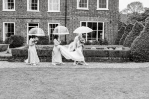 A black-and-white photo of a bride and her bridesmaids walking outside a brick building. The bride leads the way holding flowers, while two bridesmaids follow with bouquets and umbrellas, and one adjusts the bride's dress train. They are on a stone path beside a garden.
