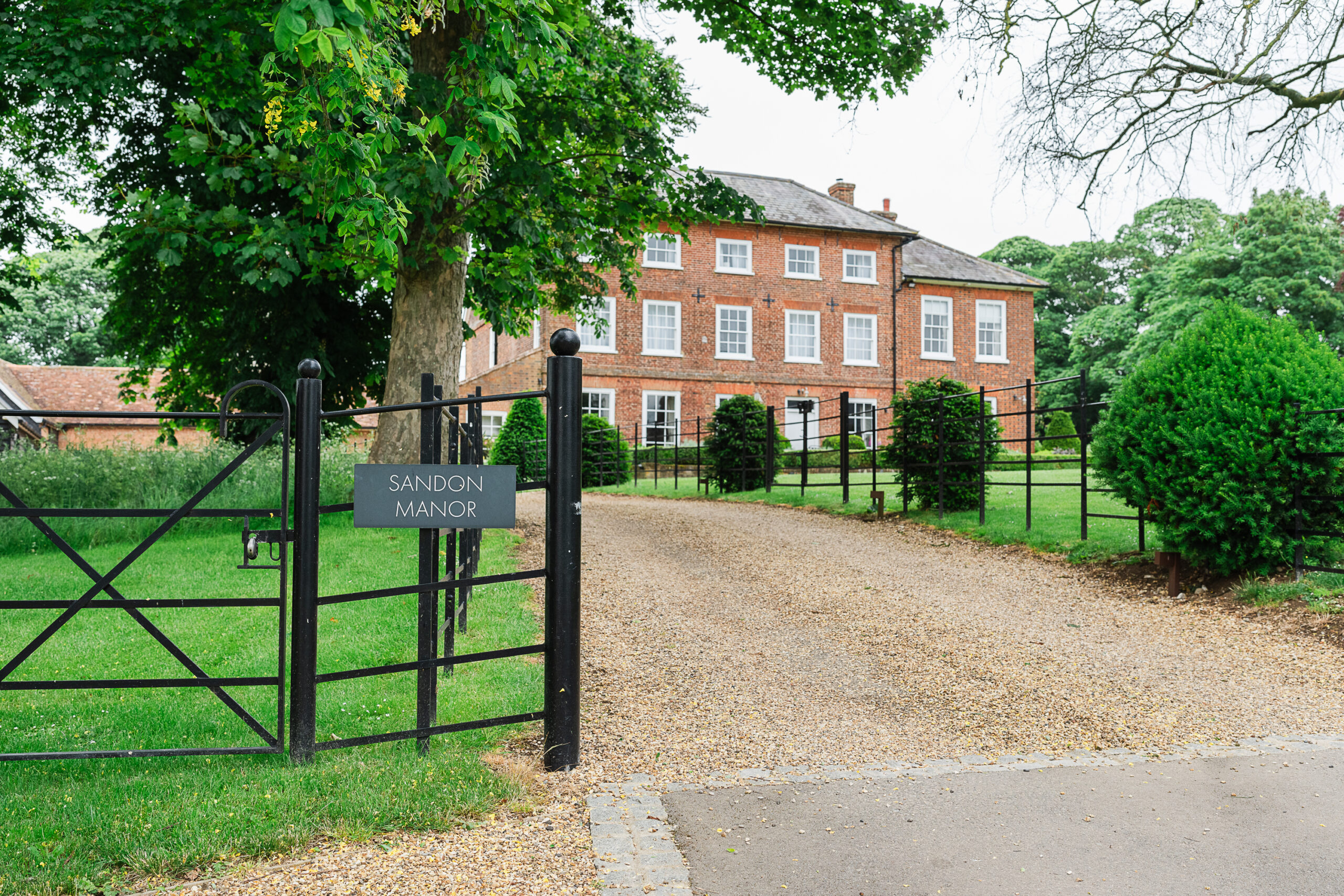 A stately, red-brick manor house, labeled "Sandon Manor," is situated at the end of a gravel driveway, bordered by manicured lawns and lush greenery. The house features large windows and is fronted by a wrought-iron gate. Trees and a well-kept hedge line the property.