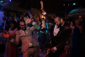 An Indian groom and his best man, enthusiastically dance the reception, expertly captured by a Hertfordshire wedding photographer. They are surrounded by their friends and family people, all smiling and enjoying the celebration. The background features festive lights and decorations, adding to the vibrant atmosphere with one of their guests applauding their dancing.