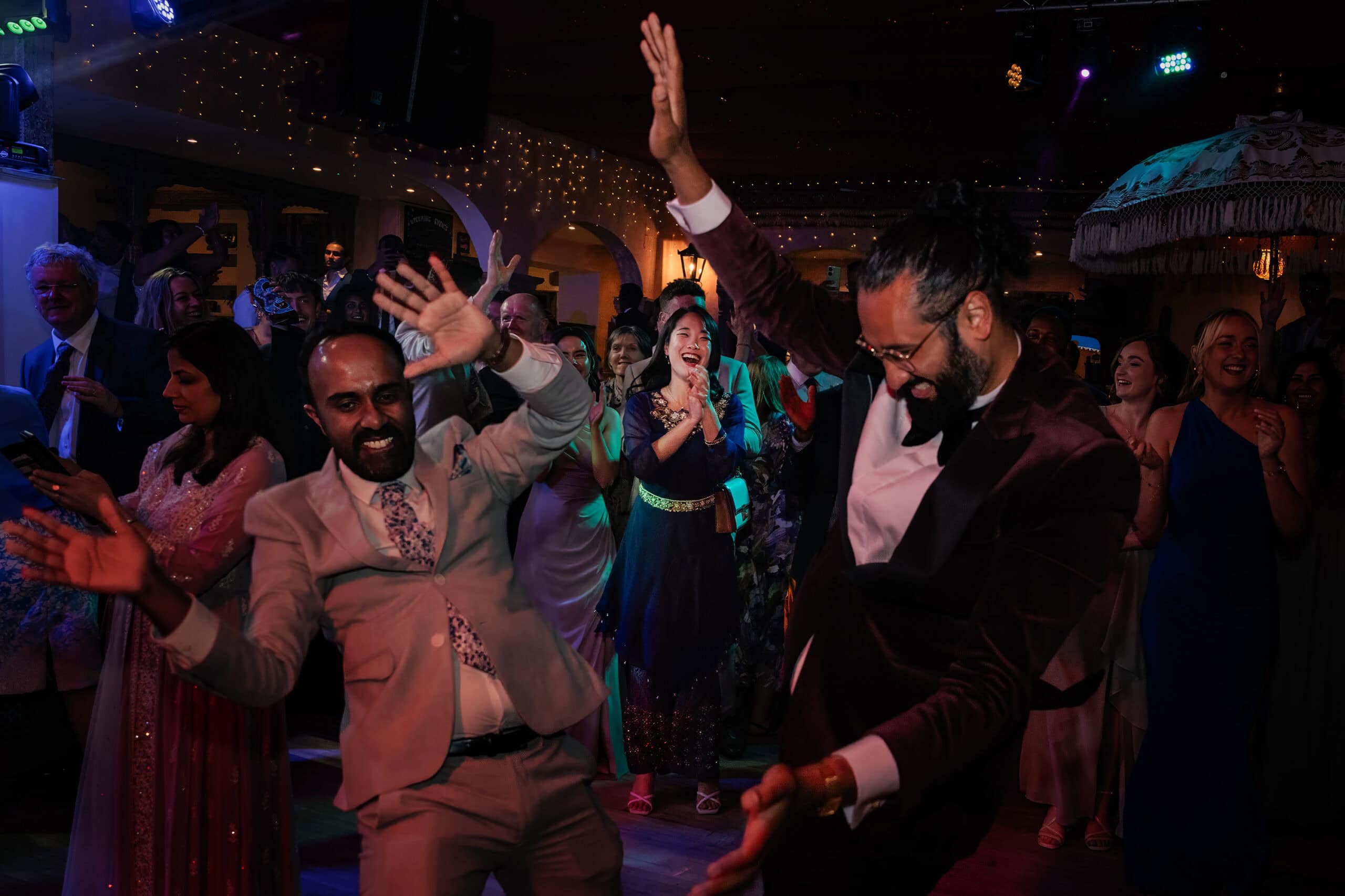 An Indian groom and his best man, enthusiastically dance the reception, expertly captured by a Hertfordshire wedding photographer. They are surrounded by their friends and family people, all smiling and enjoying the celebration. The background features festive lights and decorations, adding to the vibrant atmosphere with one of their guests applauding their dancing.