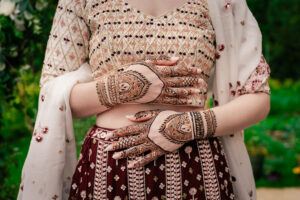A person in traditional attire at their Mehendi days before their Indian wedding, showcases intricate henna tattoo designs on their hands. They are wearing a richly patterned blouse and a maroon skirt with a white shawl draped over their shoulder, standing against a greenery-filled background.