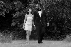 A black and white photo shows a couple walking hand in hand on a grassy meadow at Trading Boundaries wedding venue. The woman wears a white strapless dress, and the man wears a dark suit with a bow tie. Trees are visible in the background, and the couple are smiling at each other.