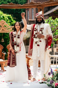 A couple stands smiling, holding hands, celebrating their wedding. The groom is dressed in traditional Indian attire adorned with red and white garlands. The bride is wearing a traditional white wedding dress. The scene is decorated with ornate wooden details and greenery in the background.