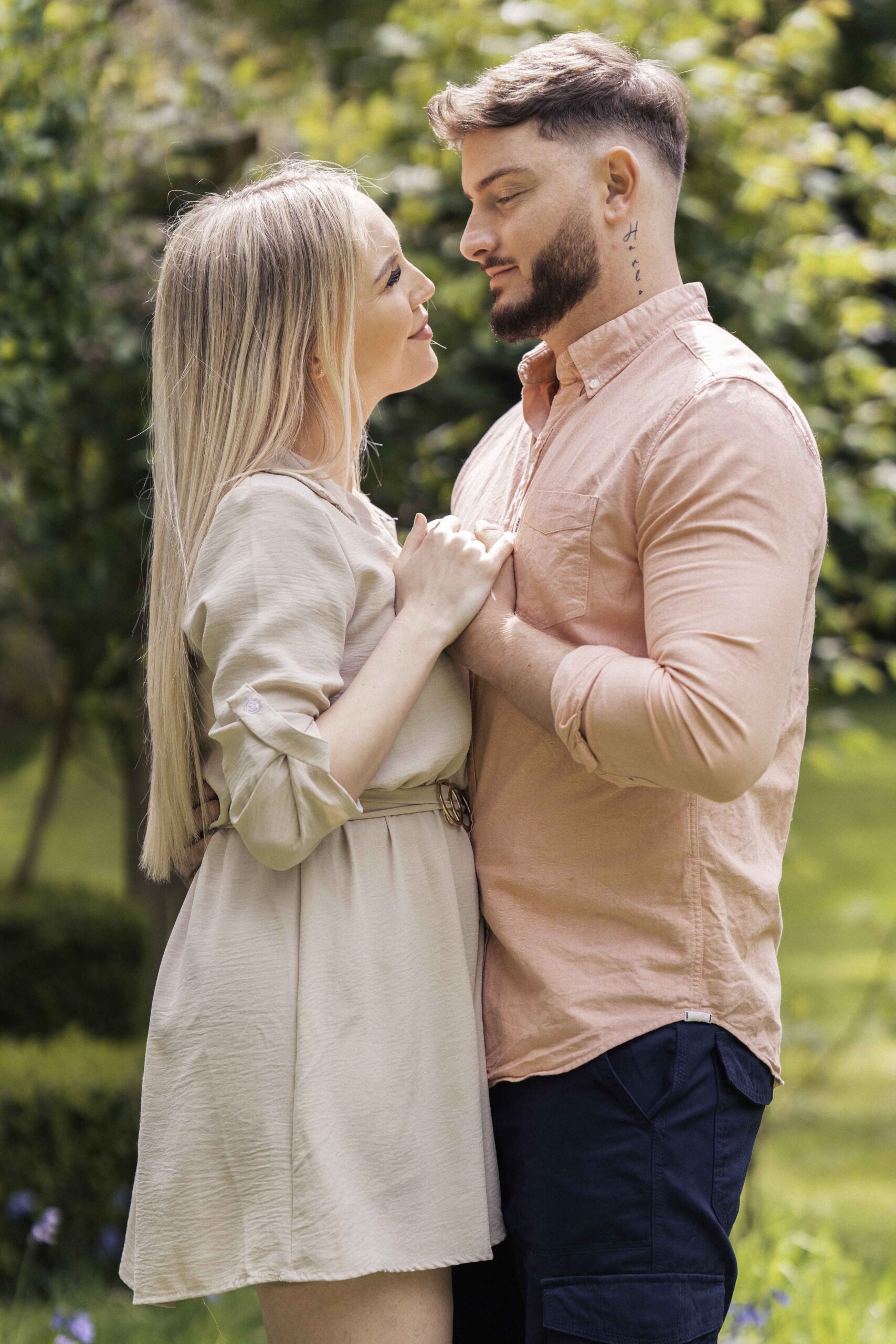 A couple stands closely in an outdoor setting, looking into each other's eyes. The woman, with long blonde hair, wears a light beige dress, while the man, with short brown hair and a beard, wears a peach-colored shirt and dark pants. They appear affectionate and connected.