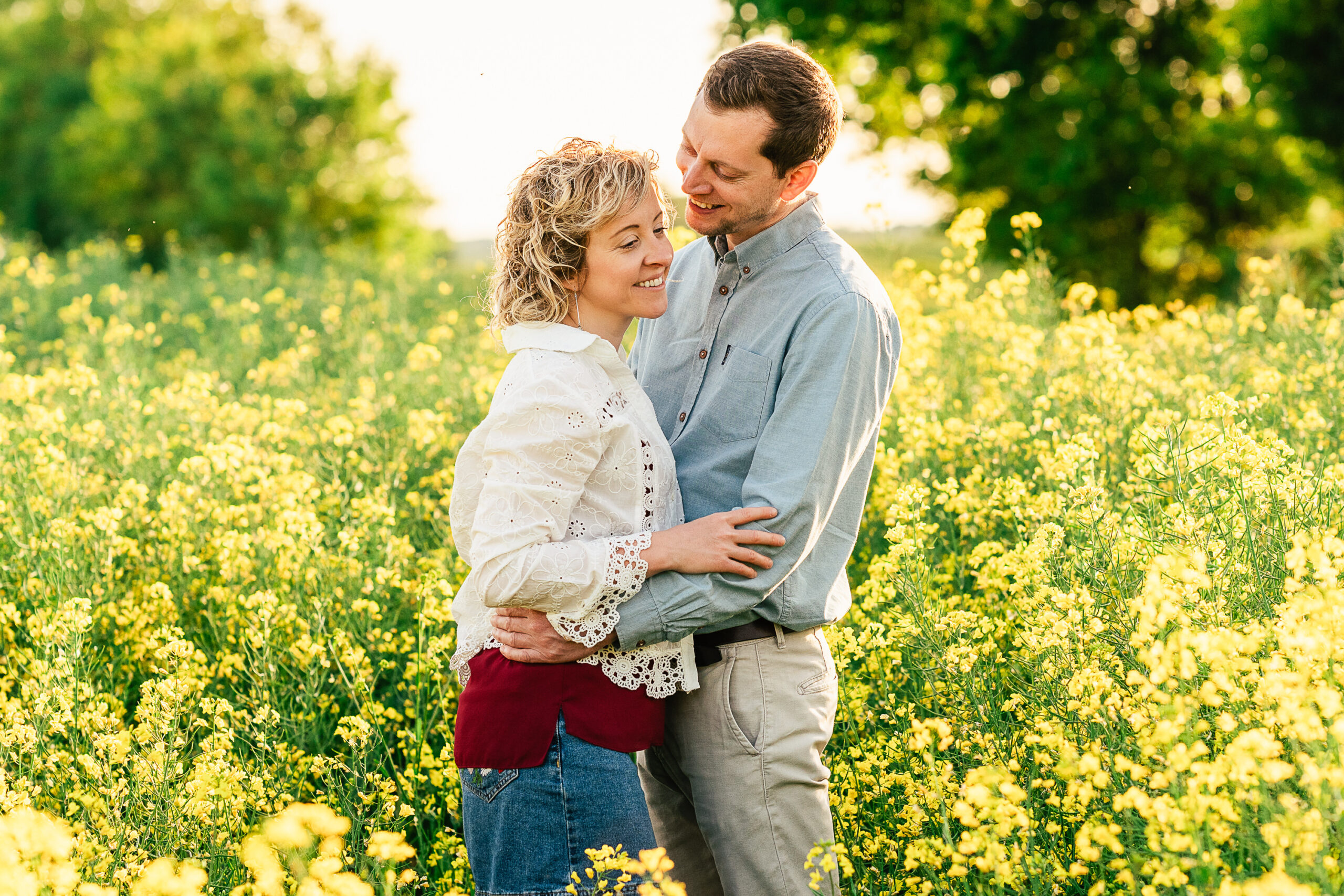 A couple stands embracing in a field of blooming yellow flowers. The woman, with curly blonde hair, wears a white lace top and denim jeans, while the man, with short brown hair, wears a blue shirt and beige pants. Both are smiling affectionately. The background is lush and green.