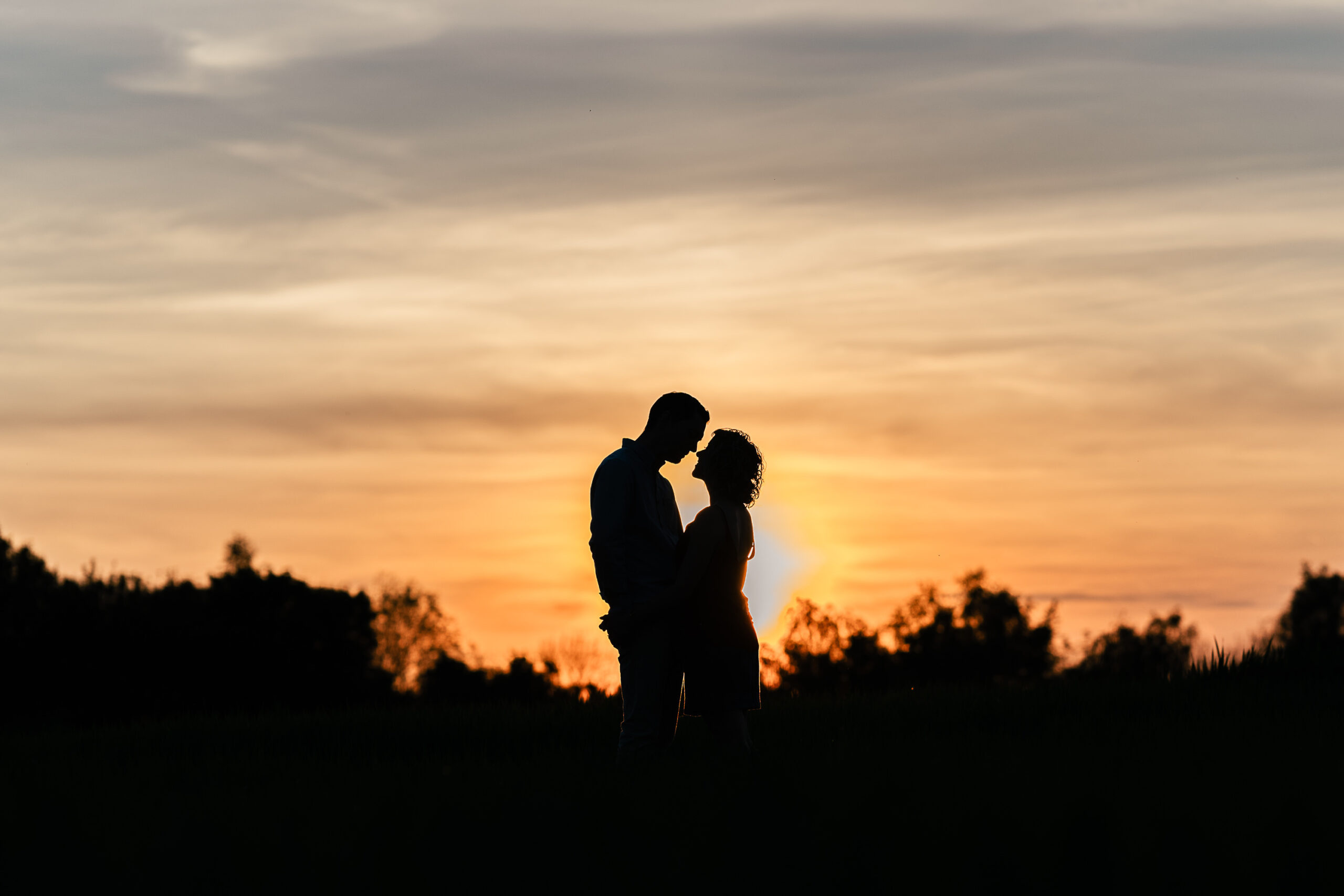 A silhouette of a couple standing close together in a field at sunset. The sky is painted with warm hues of orange and yellow as the sun sets behind them, creating a romantic and peaceful atmosphere. Trees and bushes are visible in the background.