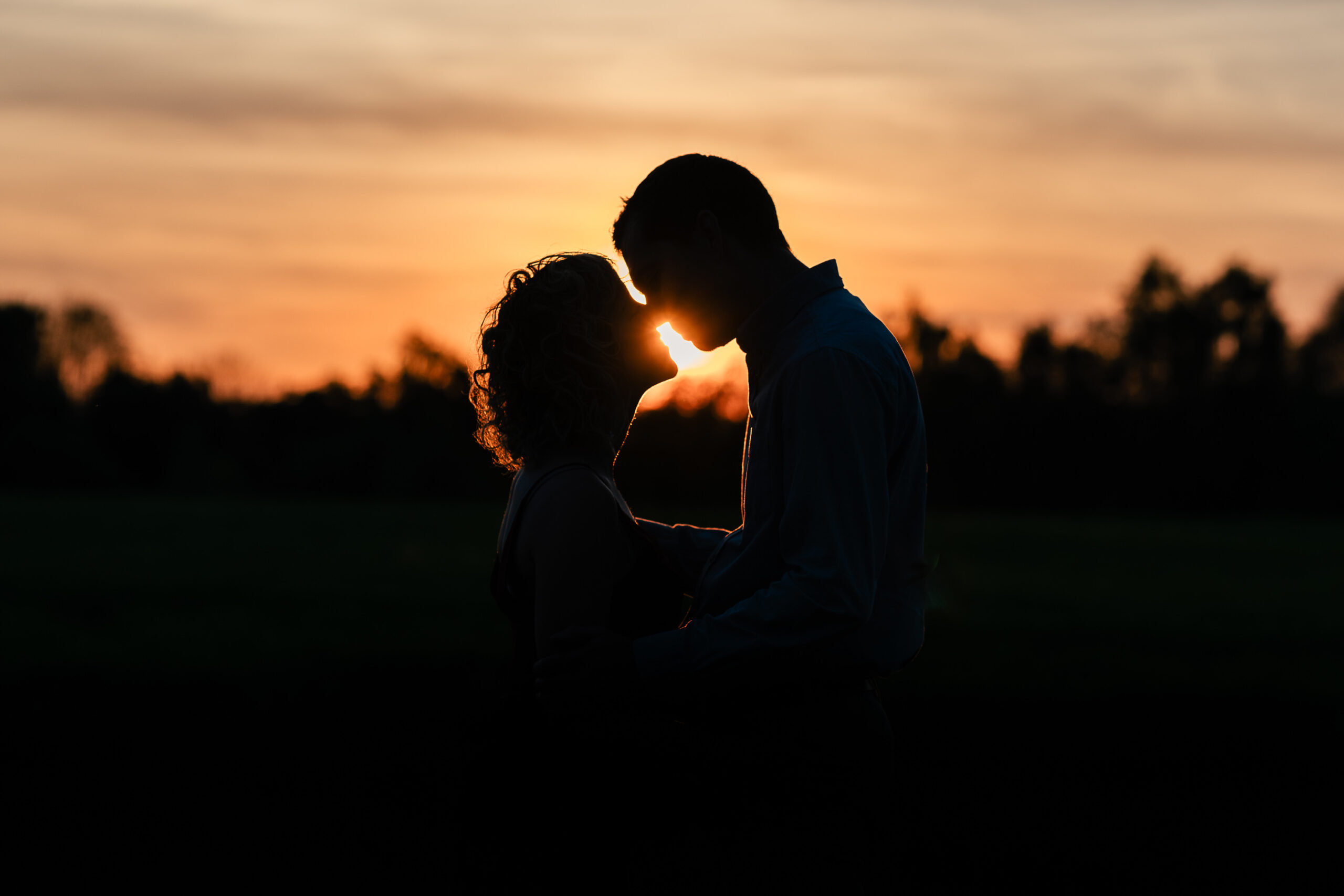 Silhouette of a couple embracing and touching foreheads against the backdrop of a vibrant sunset. The sky is tinged with orange and yellow hues, with a dark treeline in the distance. The scene exudes warmth and intimacy.