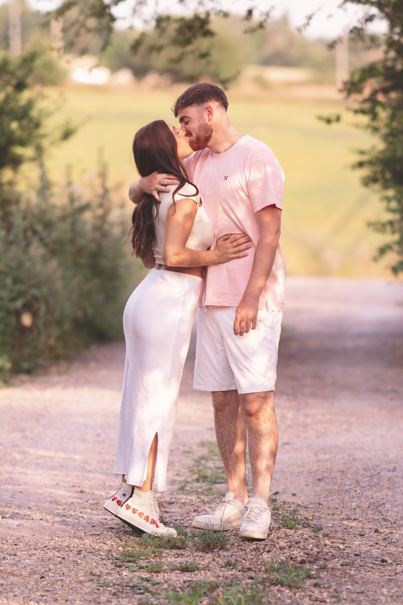 A couple embraces on a sunlit dirt path lined with trees. The woman, dressed in a white sleeveless dress and sneakers, stands on her tiptoes. The man, wearing a pink t-shirt and white shorts, leans in to kiss her forehead. The background showcases a grassy field.