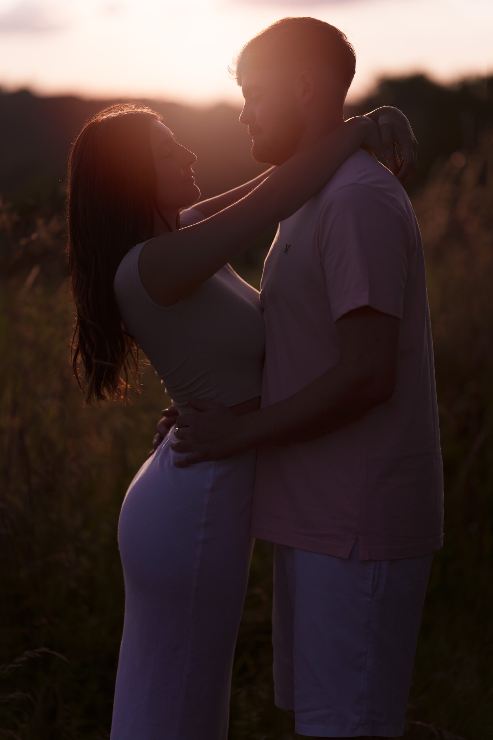 A couple stands in a field at sunset, facing each other closely. The woman has her arms around the man's neck, and he has his hands on her waist. Warm, golden light from the setting sun illuminates them, casting a romantic and intimate ambiance.