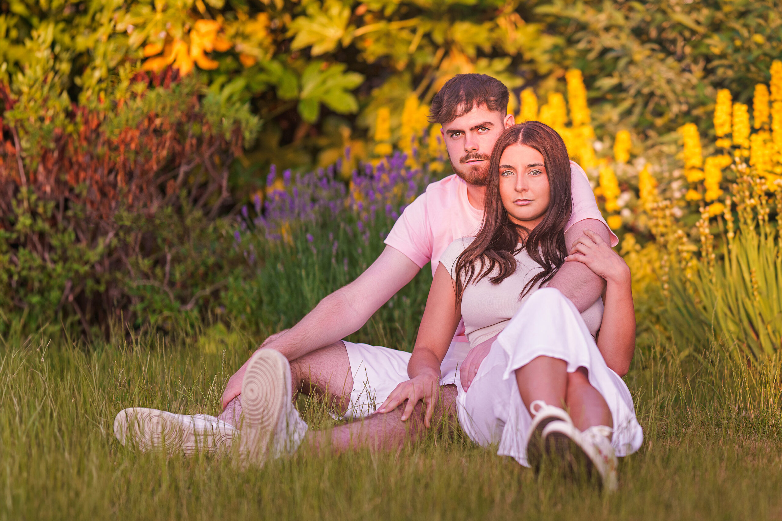 A couple sits on grass in a garden, surrounded by various flowers and green foliage. They both wear light summer clothing, with the man in a pink t-shirt and white shorts, and the woman in a white dress. The setting sun casts a warm, golden glow over the scene.