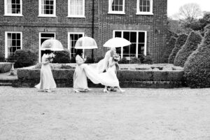 A bride in a white dress walks across a garden lawn, holding an umbrella. She is followed by three bridesmaids in long dresses, also carrying umbrellas. The background features a brick building with large windows and neatly manicured hedges.