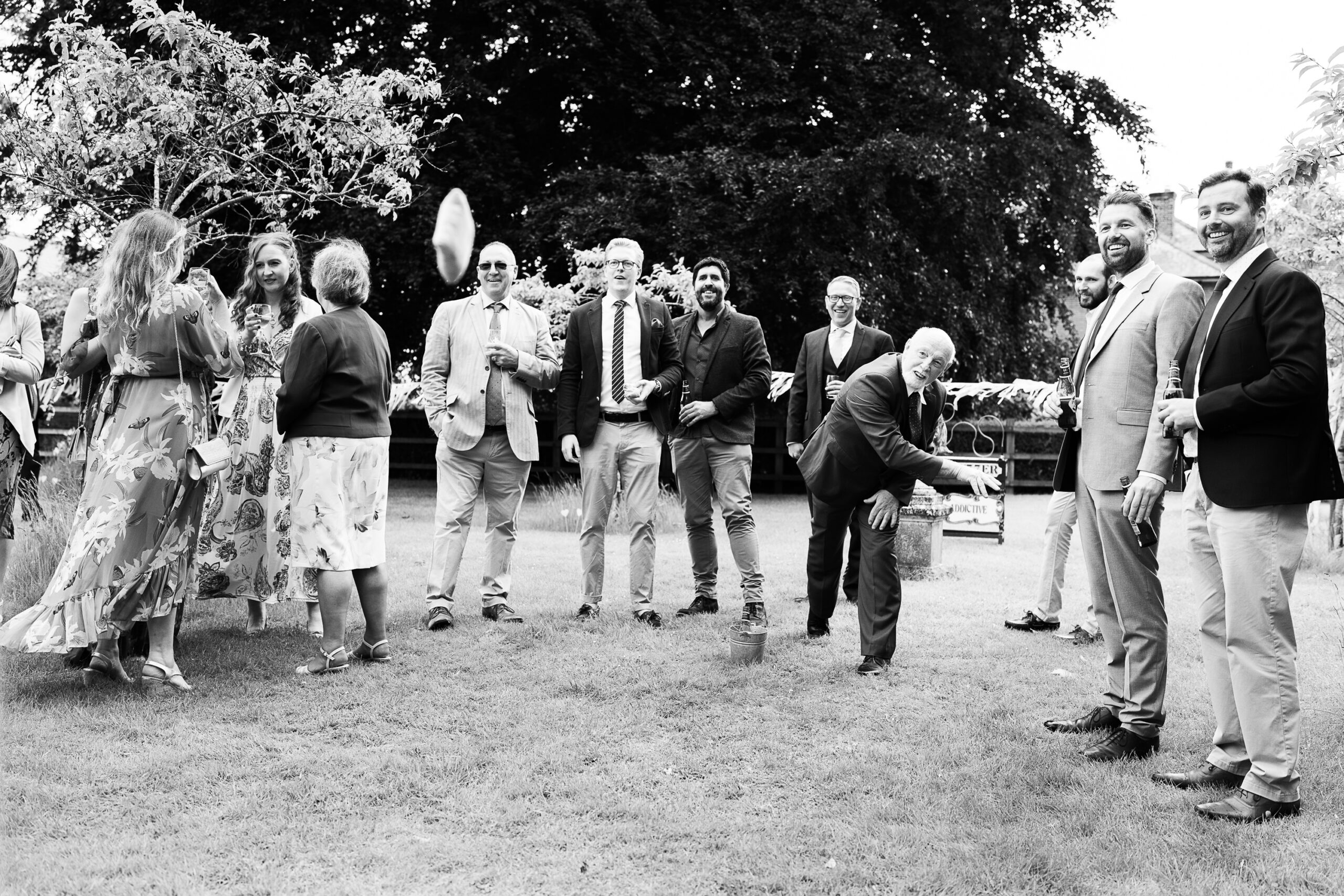 A black and white photo of people at an outdoor gathering, with some dressed formally and others more casually. One person in the center is mid-throw while several others watch in a semicircle. Trees and a wooden fence are visible in the background.