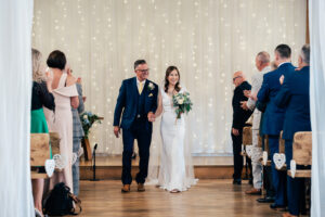 A bride in a white dress and veil walks down the aisle with the groom in a blue suit. They hold hands and smile as guests on either side applaud. The background features a curtain of twinkling lights. Wooden benches and small heart decorations are visible at Old Kent Barn, Kent.