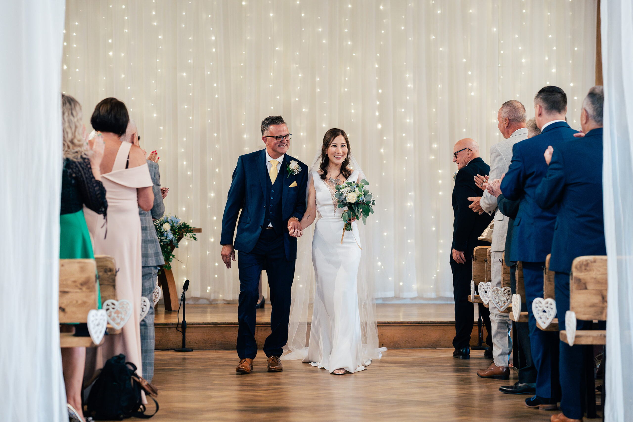 A bride in a white dress and veil walks down the aisle with the groom in a blue suit. They hold hands and smile as guests on either side applaud. The background features a curtain of twinkling lights. Wooden benches and small heart decorations are visible at Old Kent Barn, Kent.