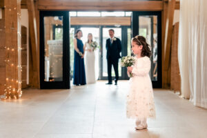 A young bridesmaid dressed in a white dress stands in the foreground holding a bouquet of flowers. In the background, the bride and one man, dressed formally, stand together, partially out of focus. The scene is at a wedding at Old Kent Barn, Kent.