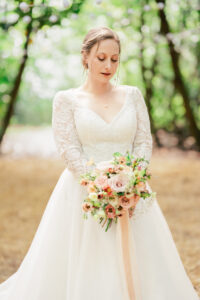 A bride stands in an outdoor wooded area, holding a bouquet of pink and white flowers. She is wearing a white, long-sleeved wedding gown with lace details and looking down toward her bouquet. The background is softly blurred with greenery and trees.