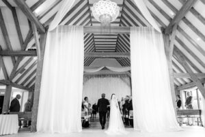 Hertfordshire wedding photographer, working in Kent, photographs a bride and her father walk down the aisle under a high, vaulted ceiling at Old Kent Barn, Kent. Two white curtains are draped on either side, and guests stand on both sides of the aisle. A large, intricate chandelier hangs from the ceiling above them. The image is in black and white.