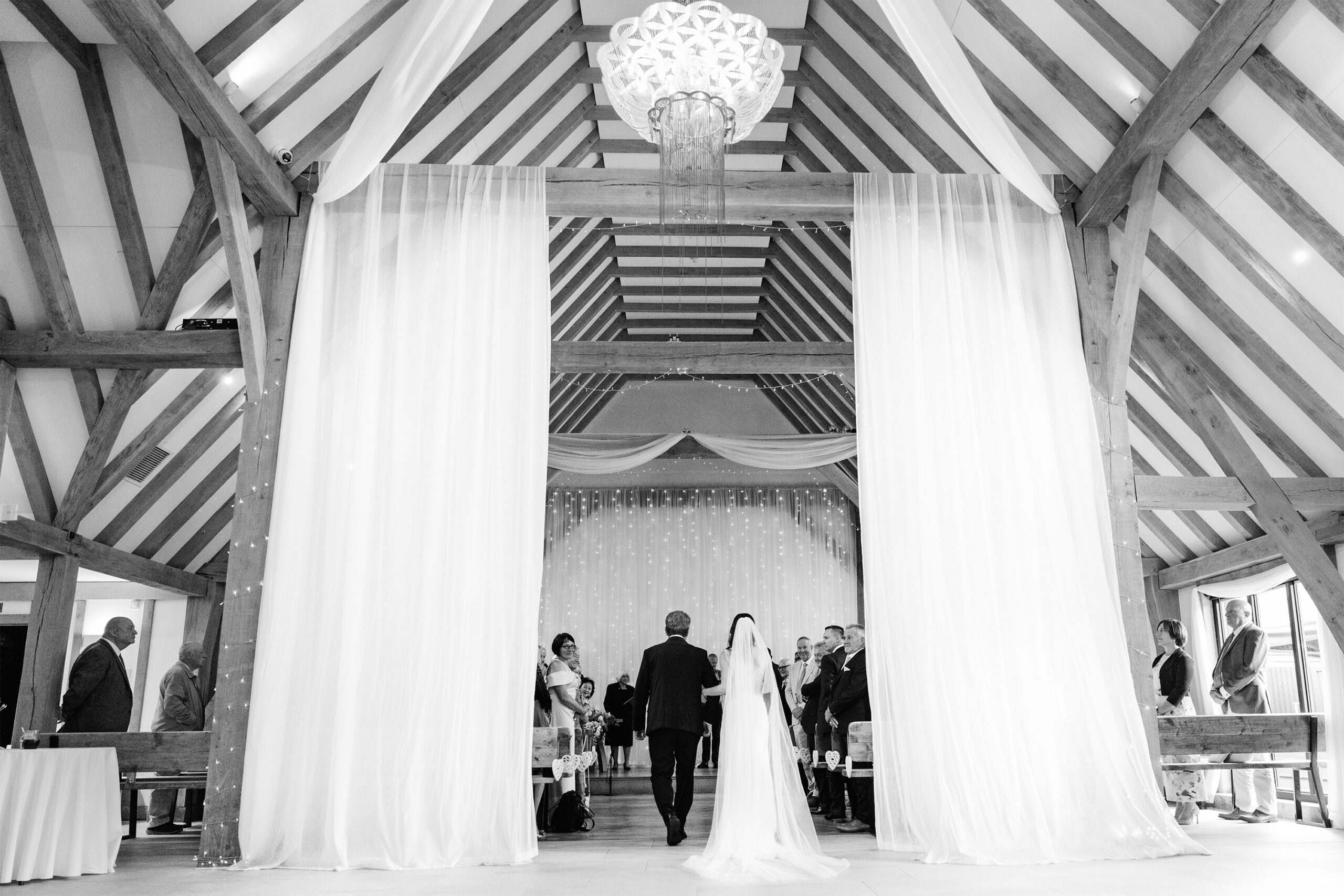 Hertfordshire wedding photographer, working in Kent, photographs a bride and her father walk down the aisle under a high, vaulted ceiling at Old Kent Barn, Kent. Two white curtains are draped on either side, and guests stand on both sides of the aisle. A large, intricate chandelier hangs from the ceiling above them. The image is in black and white.