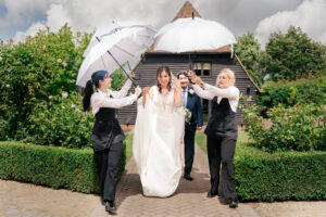 Hertfordshire wedding photographer working in Kent photographs a bride walks to the ceremony, down a garden path with her father. The bride is in a white dress. Two attendants hold umbrellas over their heads, shielding them from rain. The garden is lush and well-maintained, with a wooden building in the background at Old Kent Barn, Kent.