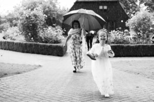 A young bridesmaid in a white dress runs on a brick path in the rain, holding her shoes in one hand and smiling. Behind her, an adult woman in a floral dress walks under an umbrella. The background shows a garden with bushes and a wooden building at Old Kent Barn, Kent.