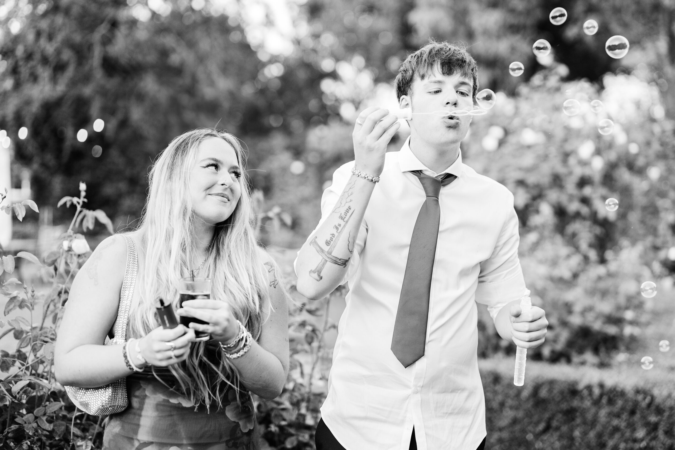 A black-and-white photo of two people outdoors. The person on the right is blowing bubbles through a bubble wand, and the person on the left is holding a drink and looking at the other with a smile. They are surrounded by greenery in a garden-like setting at Old Kent Barn, Kent.