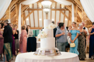 A three-tiered white wedding cake adorned with white roses is displayed on a round table with a silver stand. In the background, people are mingling in a warmly lit venue with exposed wooden beams and white drapes. At Old Kent Barn, Kent.