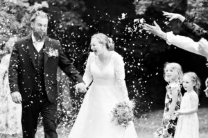 Hertfordshire wedding photographer, working in Kent, photographs a bride and groom joyfully walking hand in hand while being showered with confetti by guests. The bride holds a bouquet, laughing amidst the celebration, while the groom smiles. Young girls in floral dresses look on cheerfully. The scene is outdoors with greenery in the background.