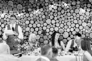 A black and white photo showing a wedding reception. A man in a vest and tie, standing and holding a microphone, reads from his speech. Four other people sit at a table, the bride and groom and the brides mother. The groom is laughing at the speech. The background has stacked logs at crown lodge, kent.