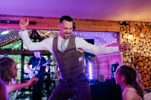 A groom in a waistcoat and tie dances enthusiastically at a lively indoor wedding event, engaging with two nearby children. Bright string lights and a wall adorned with wood slices create a festive atmosphere. A band plays a guitar in the background at crown lodge, kent.