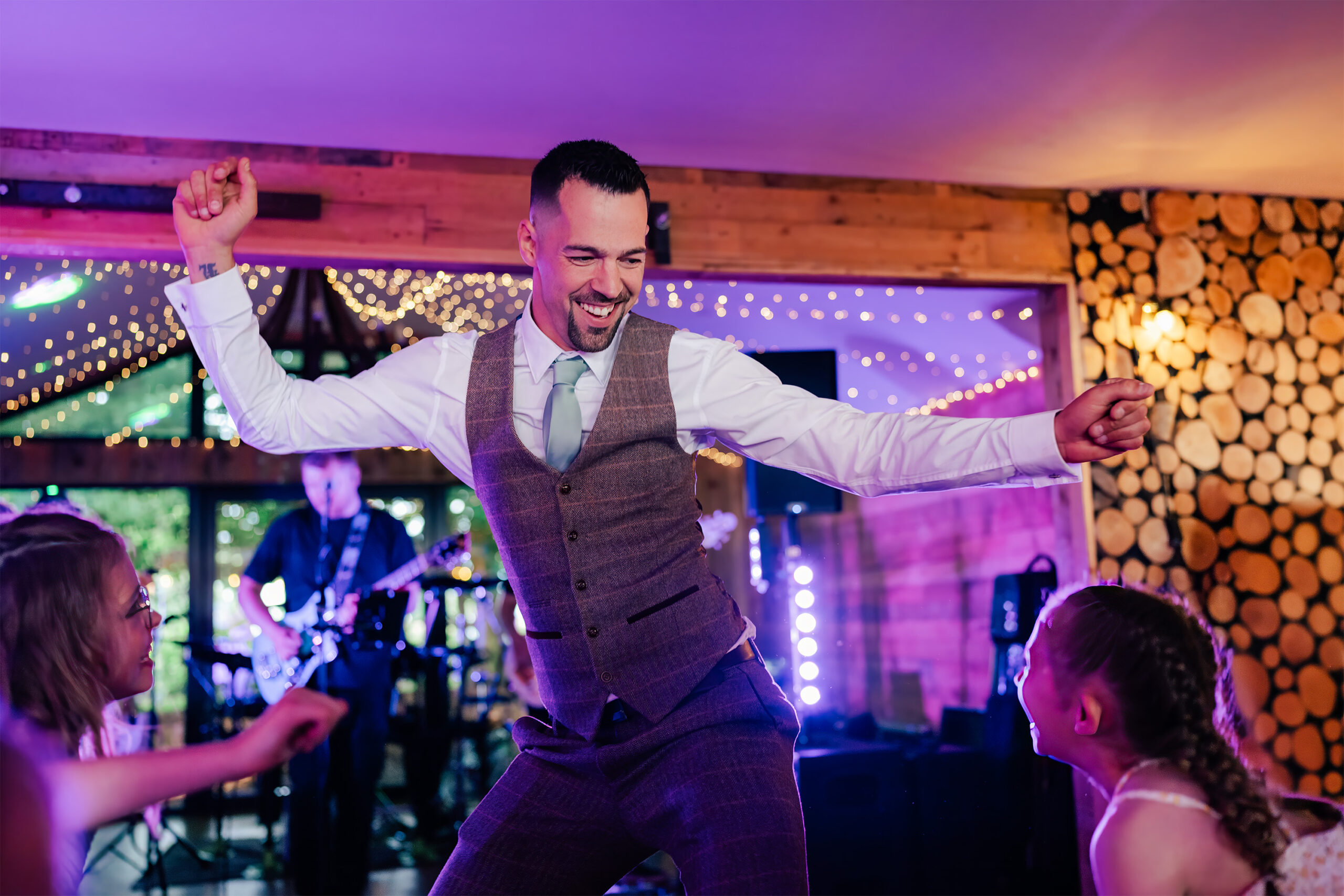 A groom in a waistcoat and tie dances enthusiastically at a lively indoor wedding event, engaging with two nearby children. Bright string lights and a wall adorned with wood slices create a festive atmosphere. A band plays a guitar in the background at crown lodge, kent.