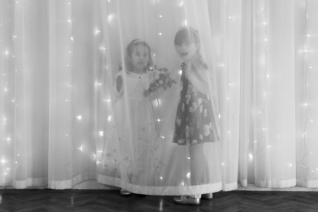 Two young children stand behind a sheer curtain adorned with tiny lights. One child holds a small bouquet of flowers, while the other smiles joyfully. Both children are dressed in light-colored clothing. The scene appears whimsical and magical at Old Kent Barn, Kent.