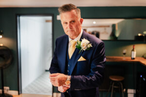 A groom in a blue suit adjusts his cufflink while standing in a modern room with green walls at Old Kent Barn, Kent. He wears a yellow tie and boutonniere, and has neatly styled hair. A wine bottle and glass sit on the counter behind him.