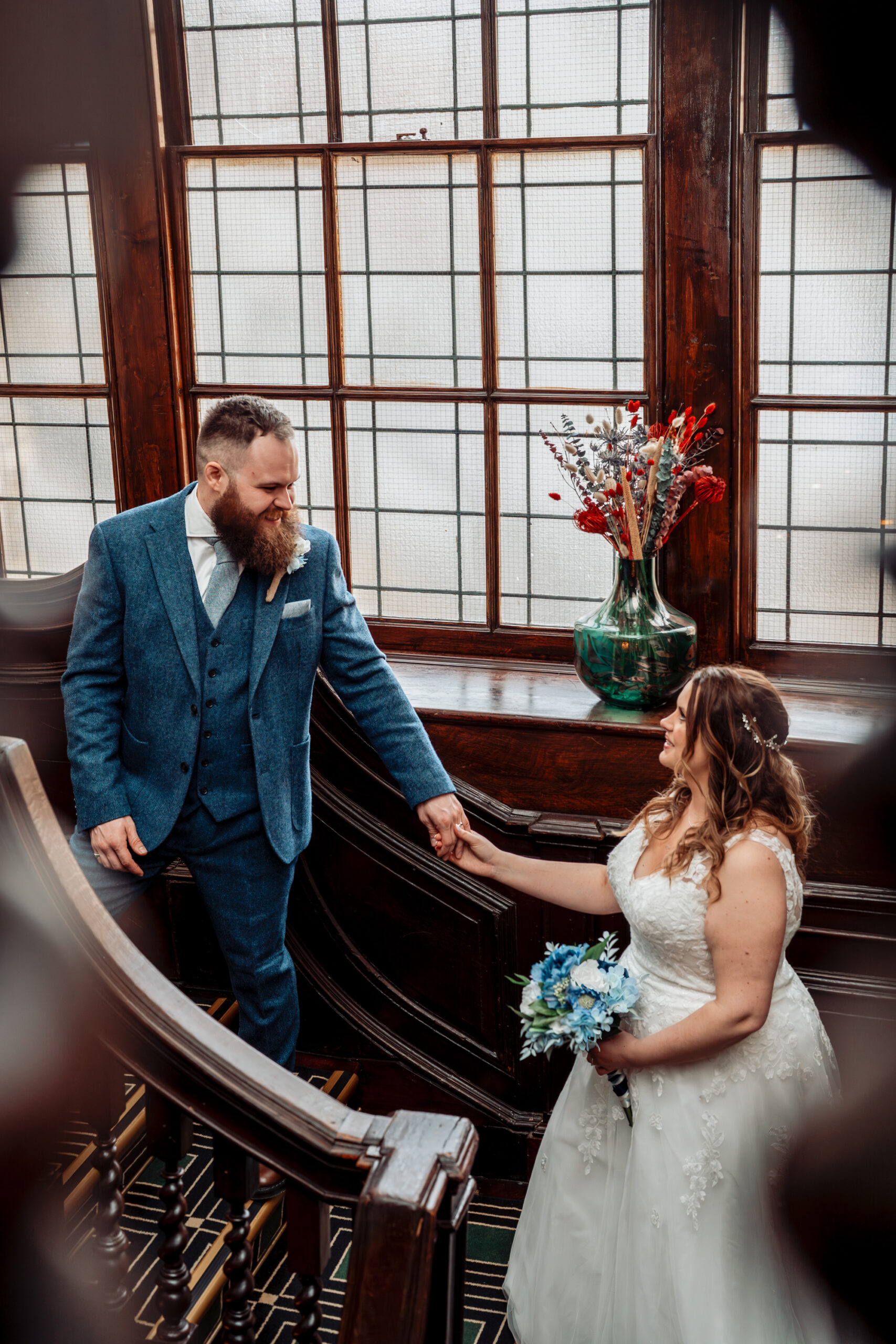 A bride with curly blonde hair stands indoors wearing a white lace wedding dress with a long train. She holds a bouquet of white and green flowers, looking down and smiling softly. The room has light green walls, tall windows with curtains, and lush greenery outside.