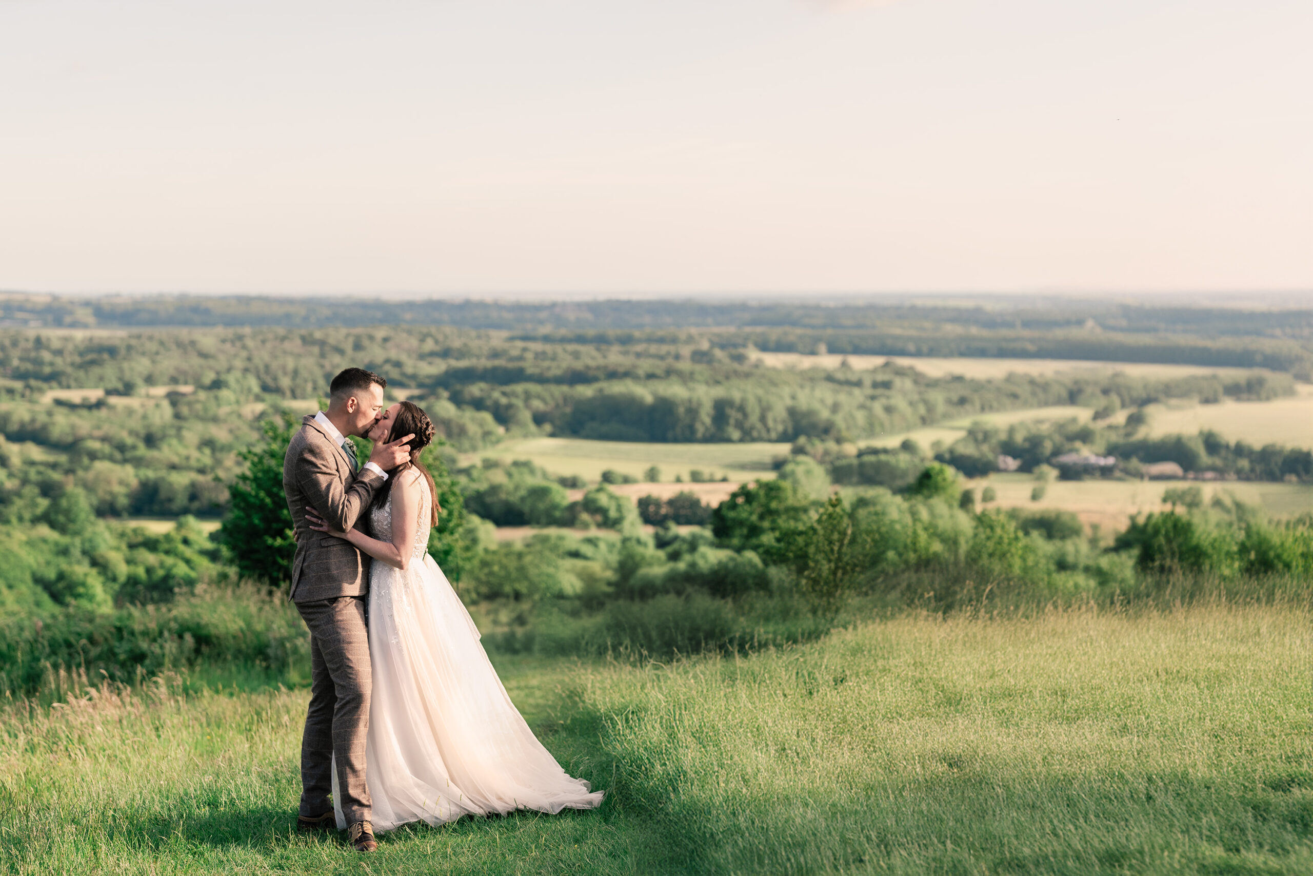 A bride in a white gown and groom in a light-colored suit share a kiss on a grassy hill, overlooking a scenic landscape of green fields and forests under a clear sky.
